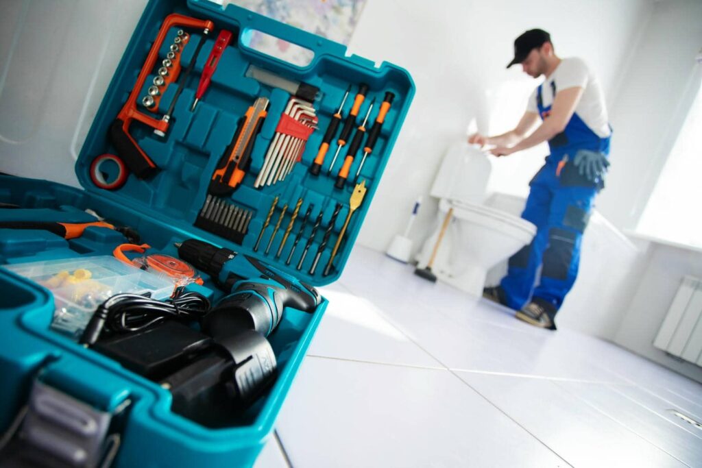 Open toolbox with various tools in the foreground and a plumber fixing a toilet in the background.