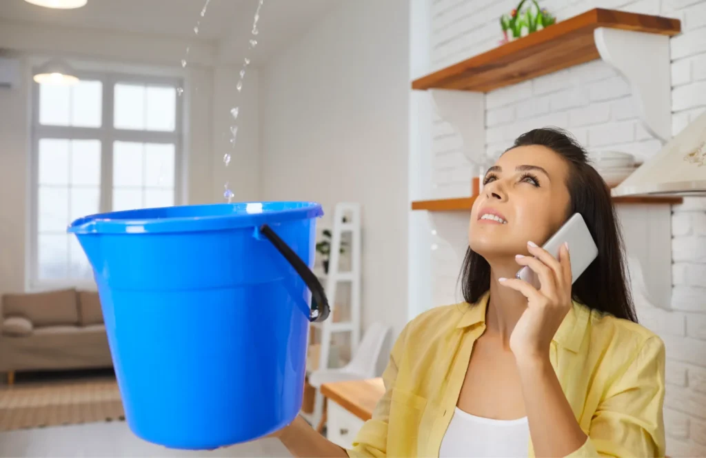 Woman holding a blue bucket to catch water leaking from the ceiling while talking on the phone.