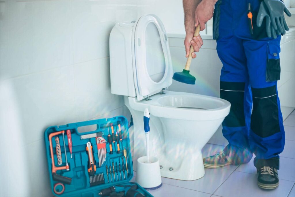 Plumber in blue pants holding a plunger next to a toilet with a tool kit on the floor.