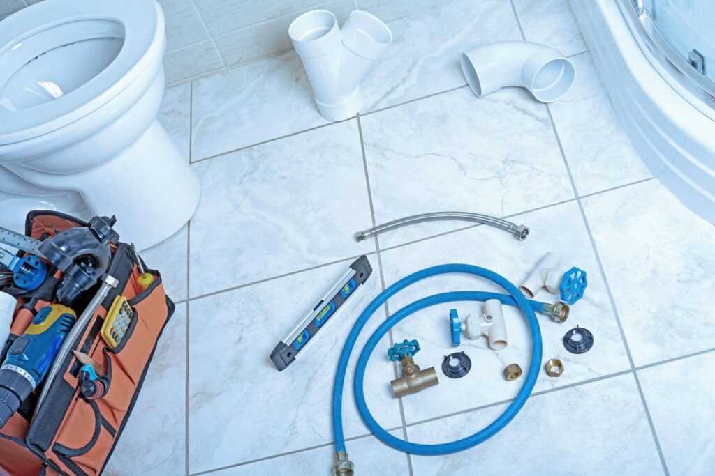 Plumbing tools and parts, including hoses, valves, and fittings, laid out on a bathroom floor near a toilet.