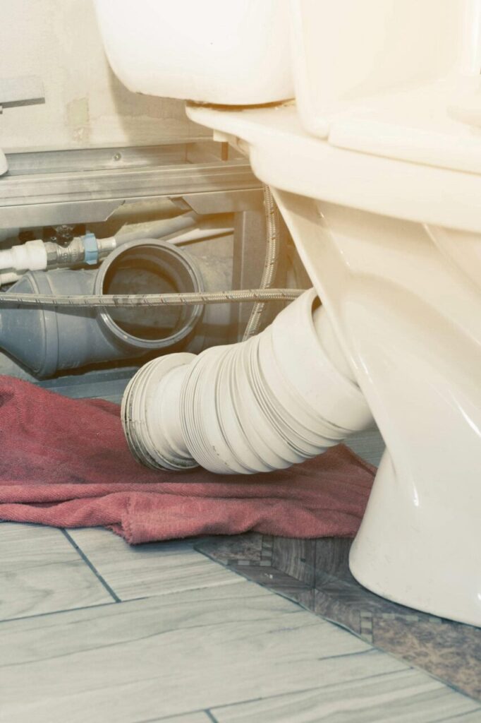 Flexible toilet drain pipe connected to a toilet with a red cloth underneath on a tiled floor.