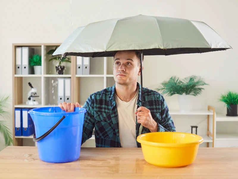 Man holding an umbrella indoors with water dripping into a blue bucket on a table. leak detection services